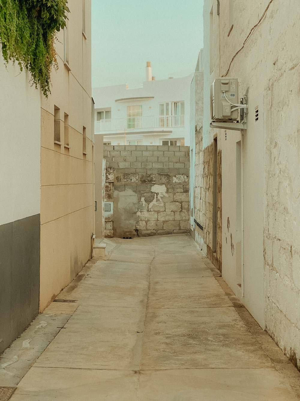 A narrow alleyway between two buildings with beige and white walls. The alleyway has a paved floor and ends at a partially bricked-up wall with an unfinished look. There are a few windows, an air conditioning unit mounted on the right wall.