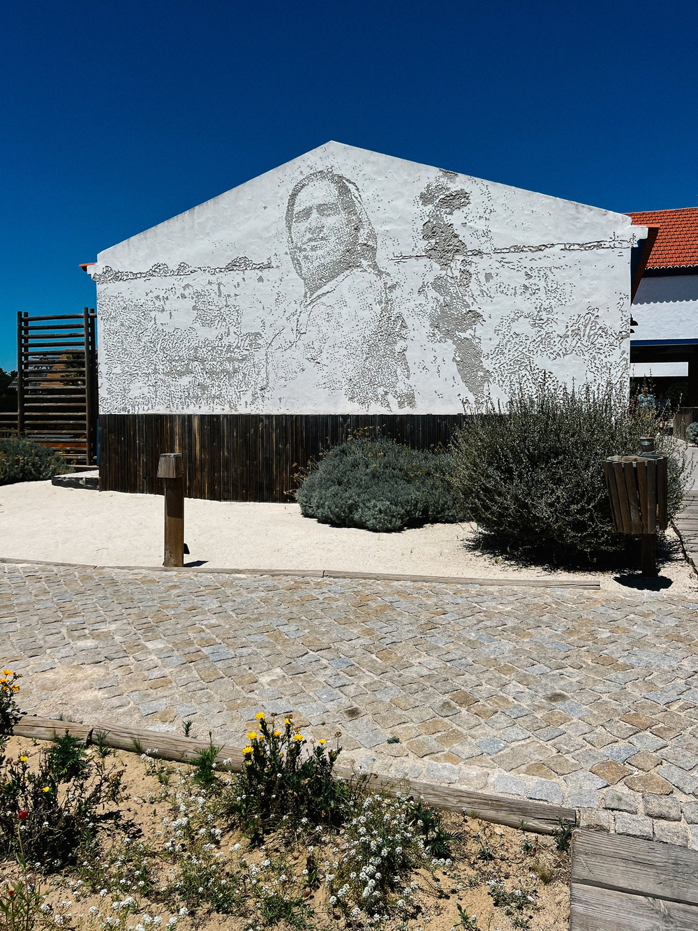 A white building with a mural depicting a human figure in a textured, dotted style. The building sits in a landscaped area with cobblestone pathways, sparse greenery, and a clear blue sky overhead.