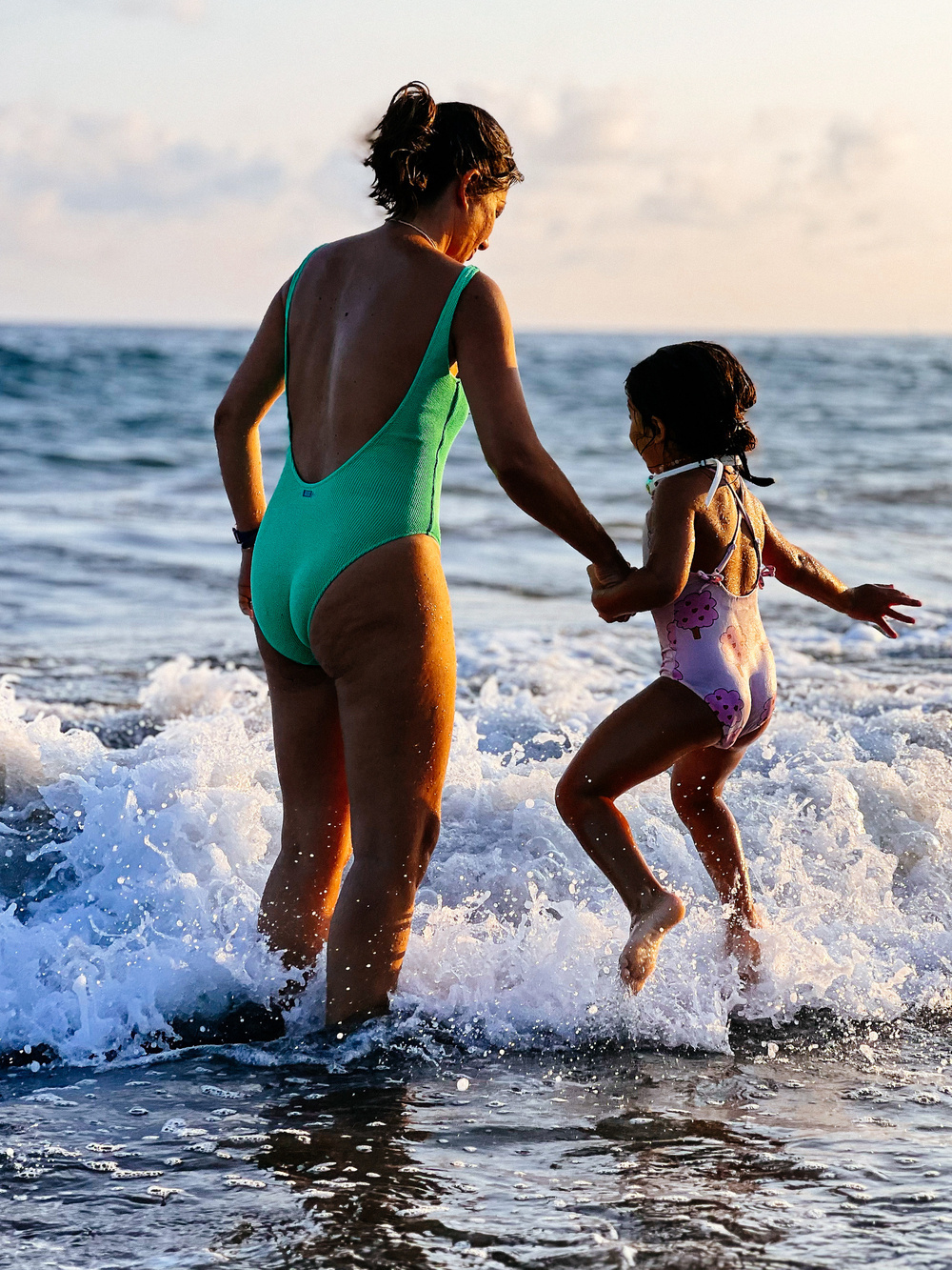 A woman and a young girl holding hands and playing in the ocean waves at the beach. The woman is wearing a green swimsuit, and the girl is wearing a purple swimsuit with a floral pattern. They are backlit by the setting sun.