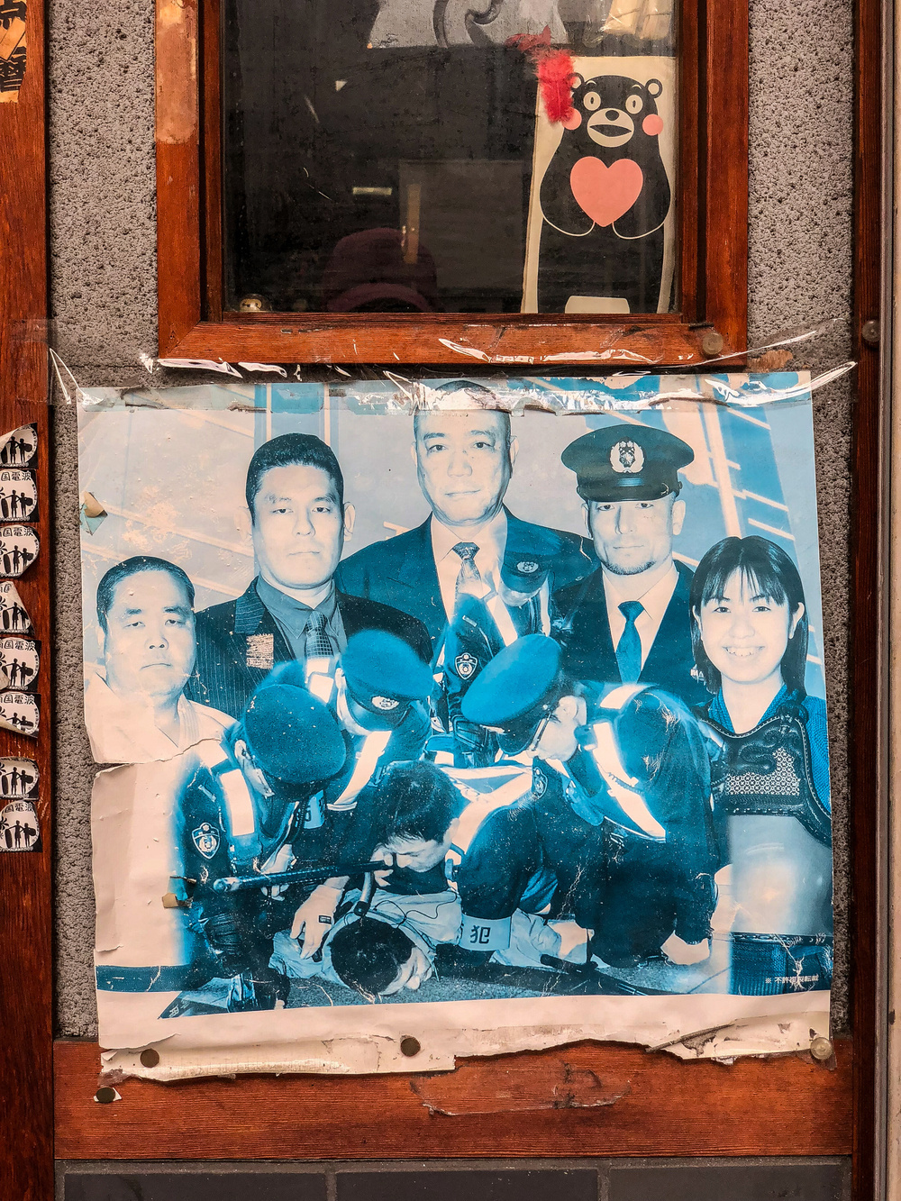 A weathered poster featuring several people, some in professional or formal attire and others in uniform, affixed to a window with a reflection and a cartoon bear illustration visible in the background.