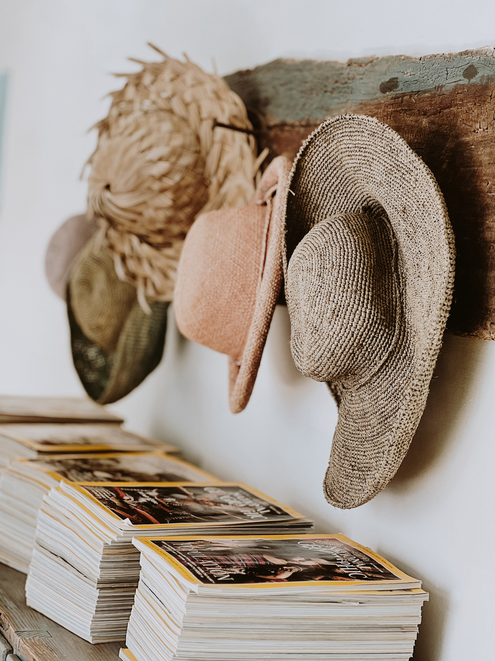 The image shows several straw hats hanging on a rustic wooden peg rack mounted on a wall. Below the hats, there are neatly stacked magazines with yellow borders and various cover images.