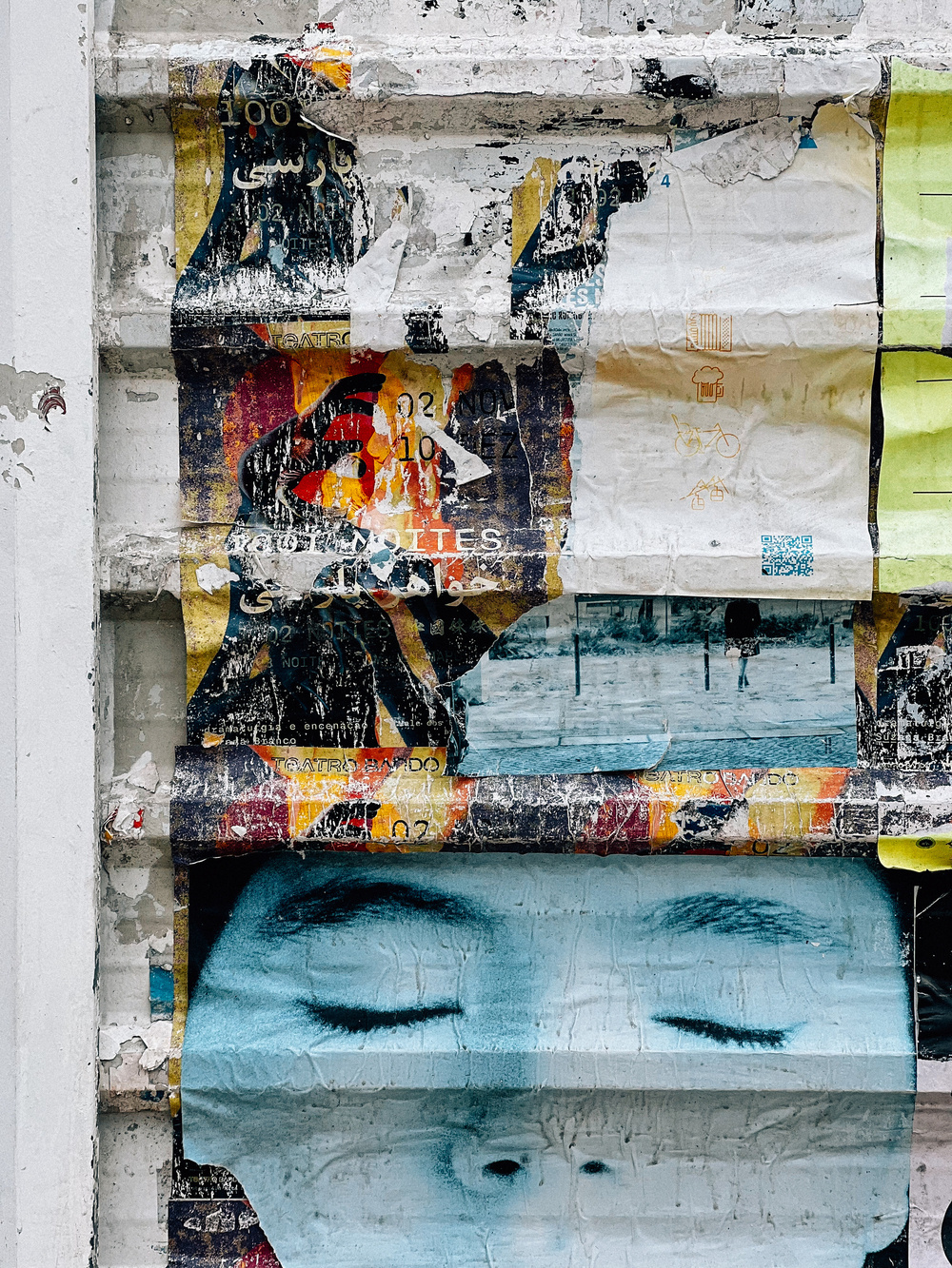A wall covered in layers of torn and weathered posters and street art. The prominent feature is a large image of a person’s closed eyes in blue hues at the bottom. Other elements include partially ripped posters.