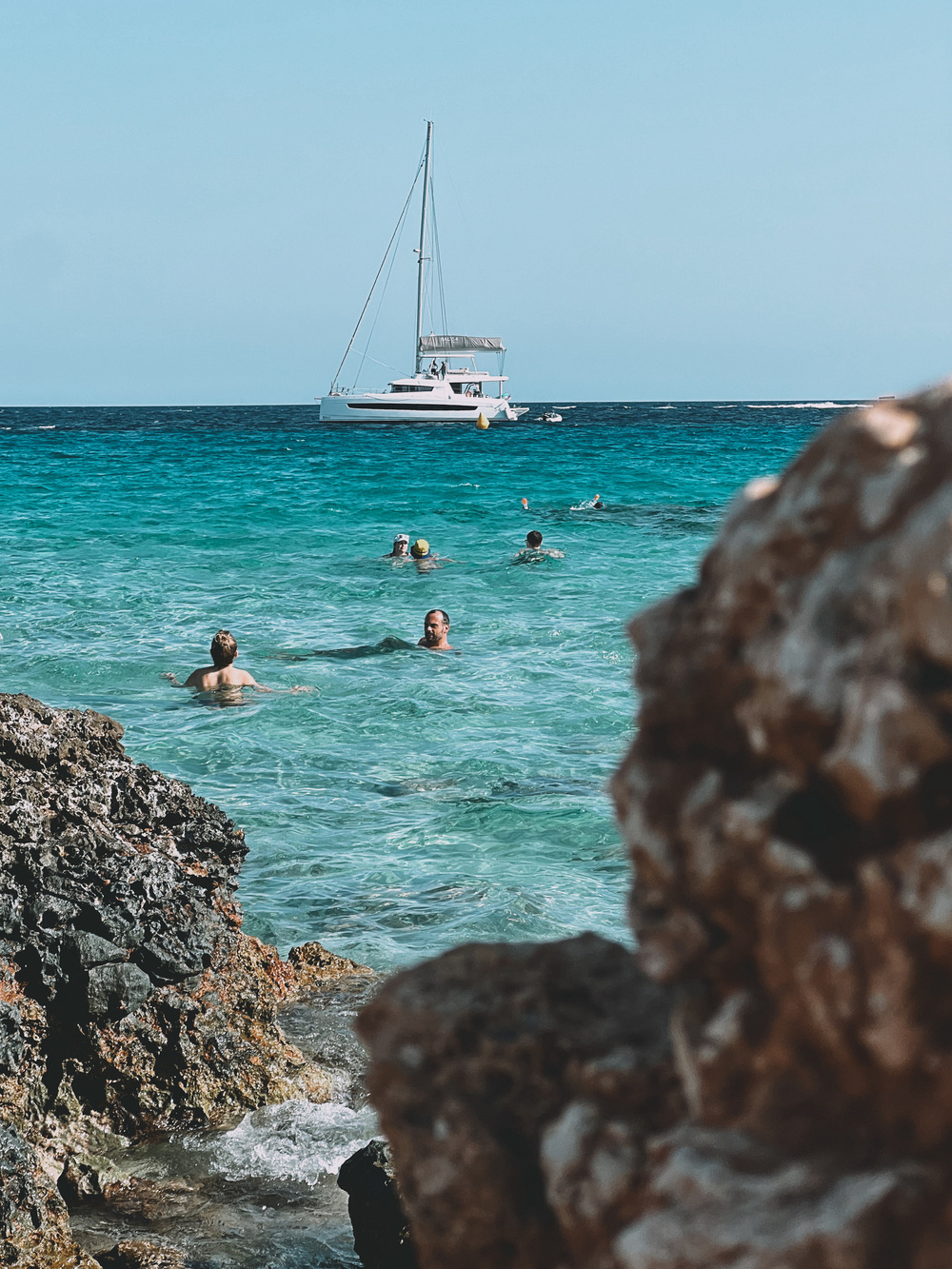 People swimming in clear blue water near rocky shore, with a white sailboat anchored in the background.
