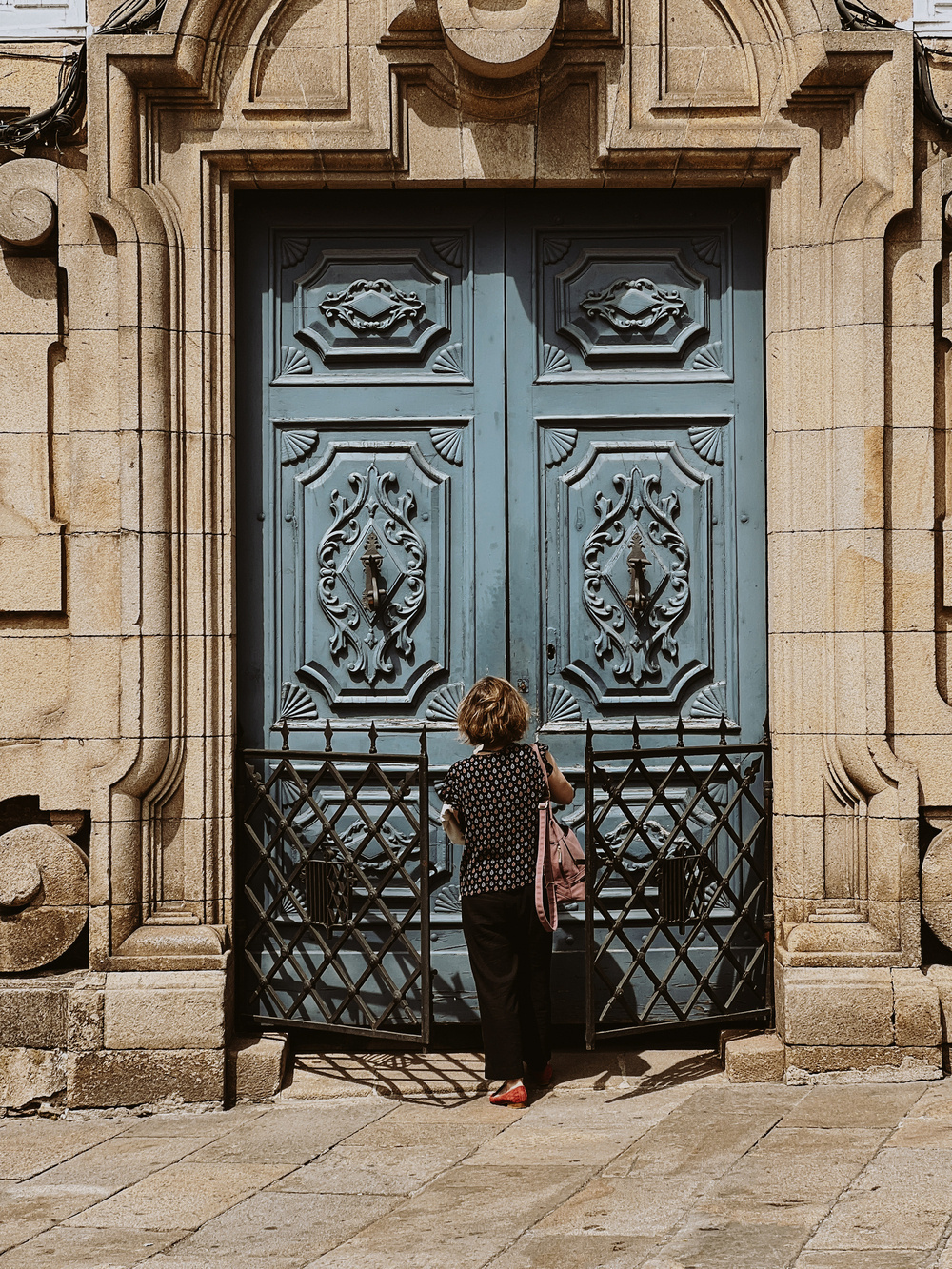 A person stands in front of a large, ornate blue door, reaching towards it. The door is framed by intricate stonework and features elaborate decorative elements. The person is holding a pink bag and is partly enclosed by a black wrought iron gate.