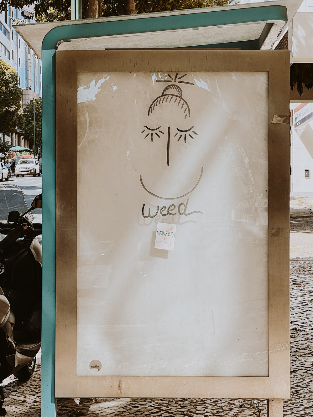 An outdoor billboard features a simple, stylized drawing of a smiling face with closed eyes and a bun. Below the drawing is the word &ldquo;weed,&rdquo; and a small piece of paper is attached with the words &ldquo;NATUREZA VOLTA.&quot;