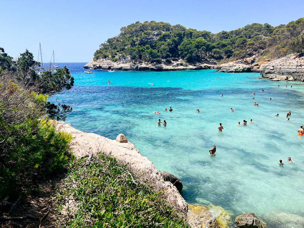 Pristine beach scene with clear turquoise water and people swimming. The coastline is surrounded by lush greenery and rocky cliffs. Sailboats are anchored in the distance under a clear blue sky.