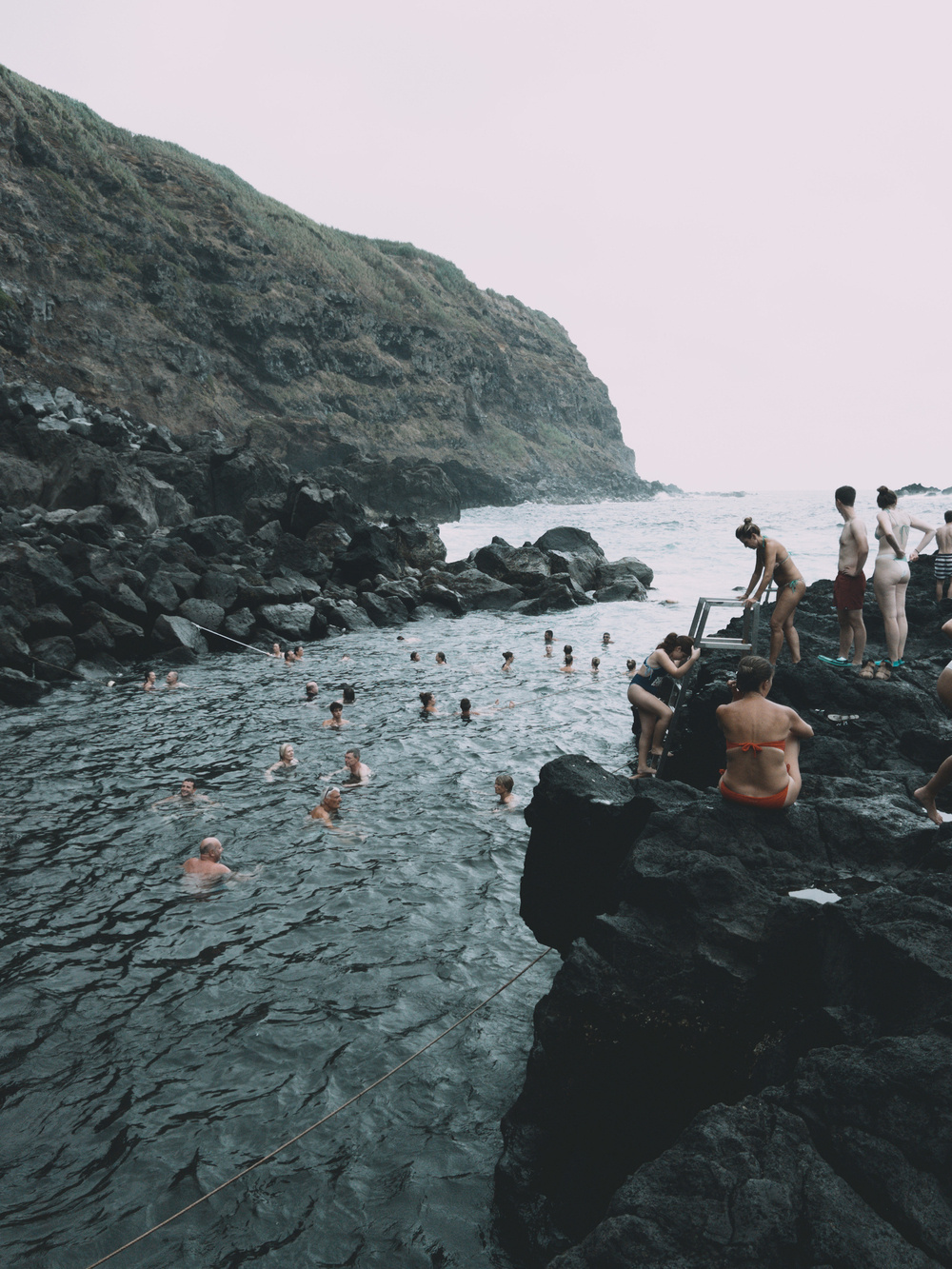 People swimming and relaxing in a natural rocky pool alongside a rugged coastline. Some individuals are entering or exiting the water using a ladder, while others are sitting or standing on the surrounding rocks. The water is choppy, and the backdrop features steep, rocky, cliffs. 