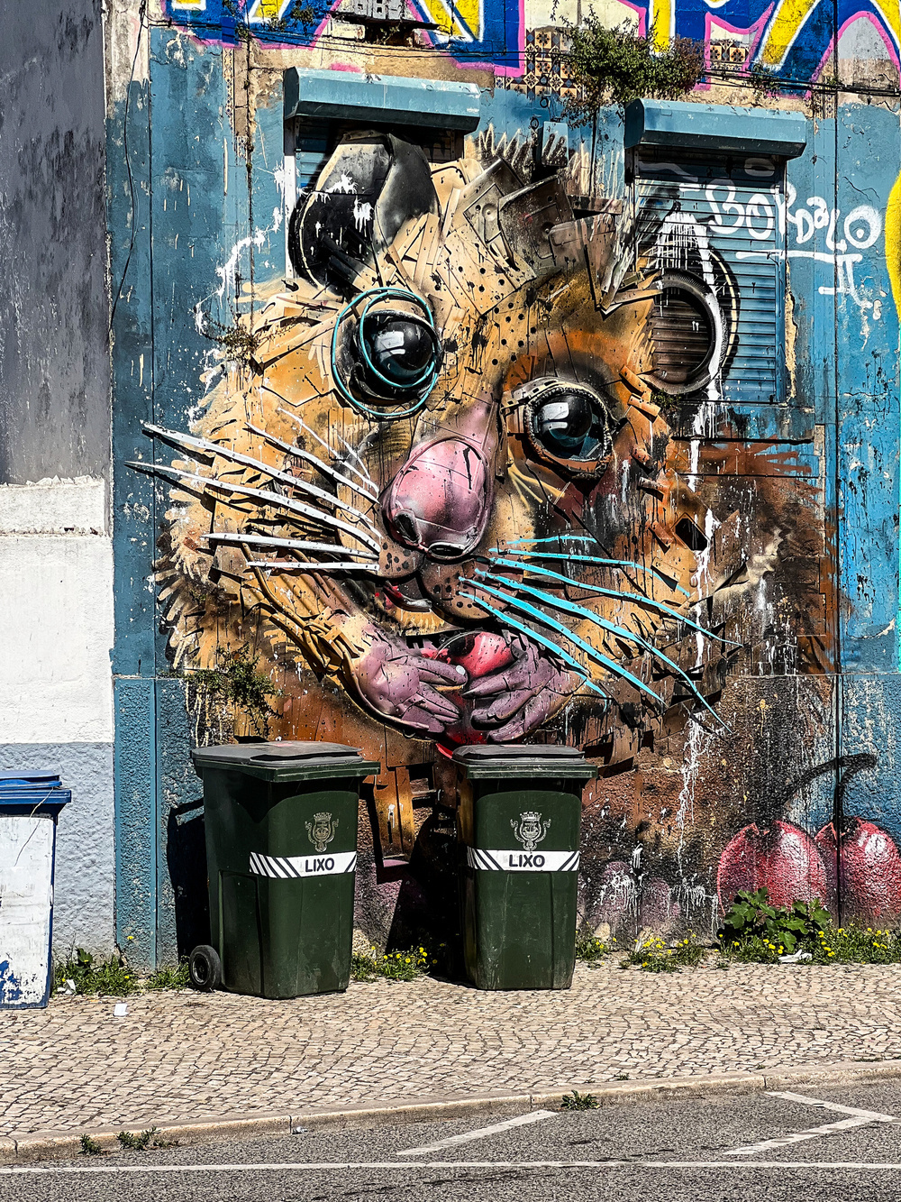 A vibrant mural of a large, colorful rodent holding an object with both hands, painted against a building facade. Two green trash bins in front of the mural are labeled &ldquo;LIXO,&rdquo; the Portuguese word for garbage.