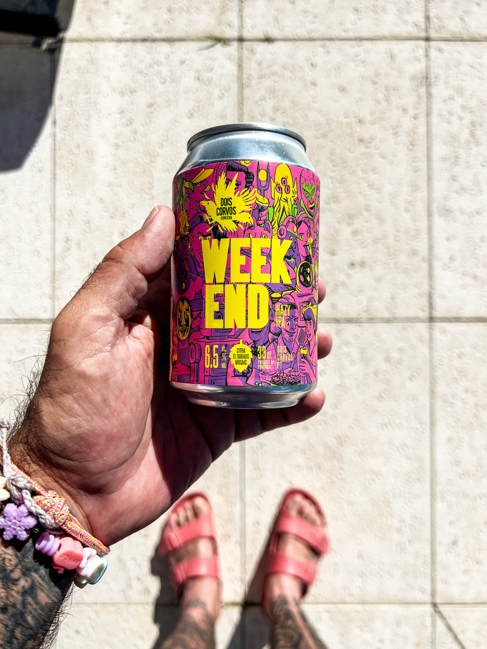 A can of “Weekend Hazy IPA” from Dois Corvos Brewery is held by a person with visible tattoos and wearing a multicolored bracelet. The can features colorful, artistic graphics with a yellow logo and details about the beer.