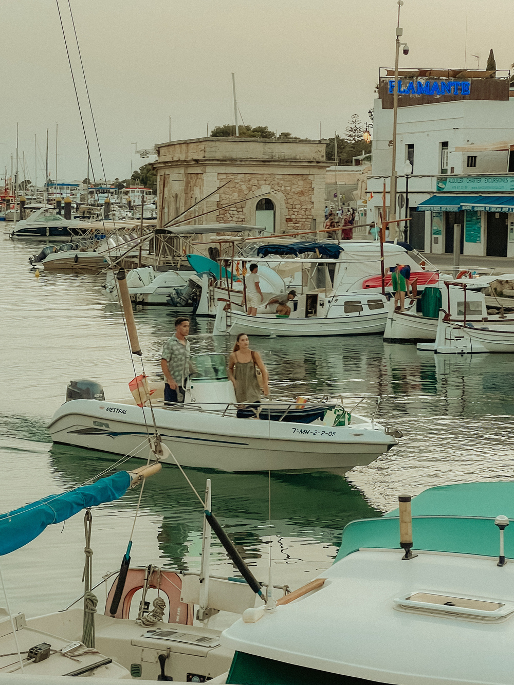 A serene marina scene with several docked boats and a small motorboat carrying two individuals. The background includes stone buildings and a neon sign reading &ldquo;FLAMANTE.&rdquo; The water is calm, with reflections of the boats and buildings visible.