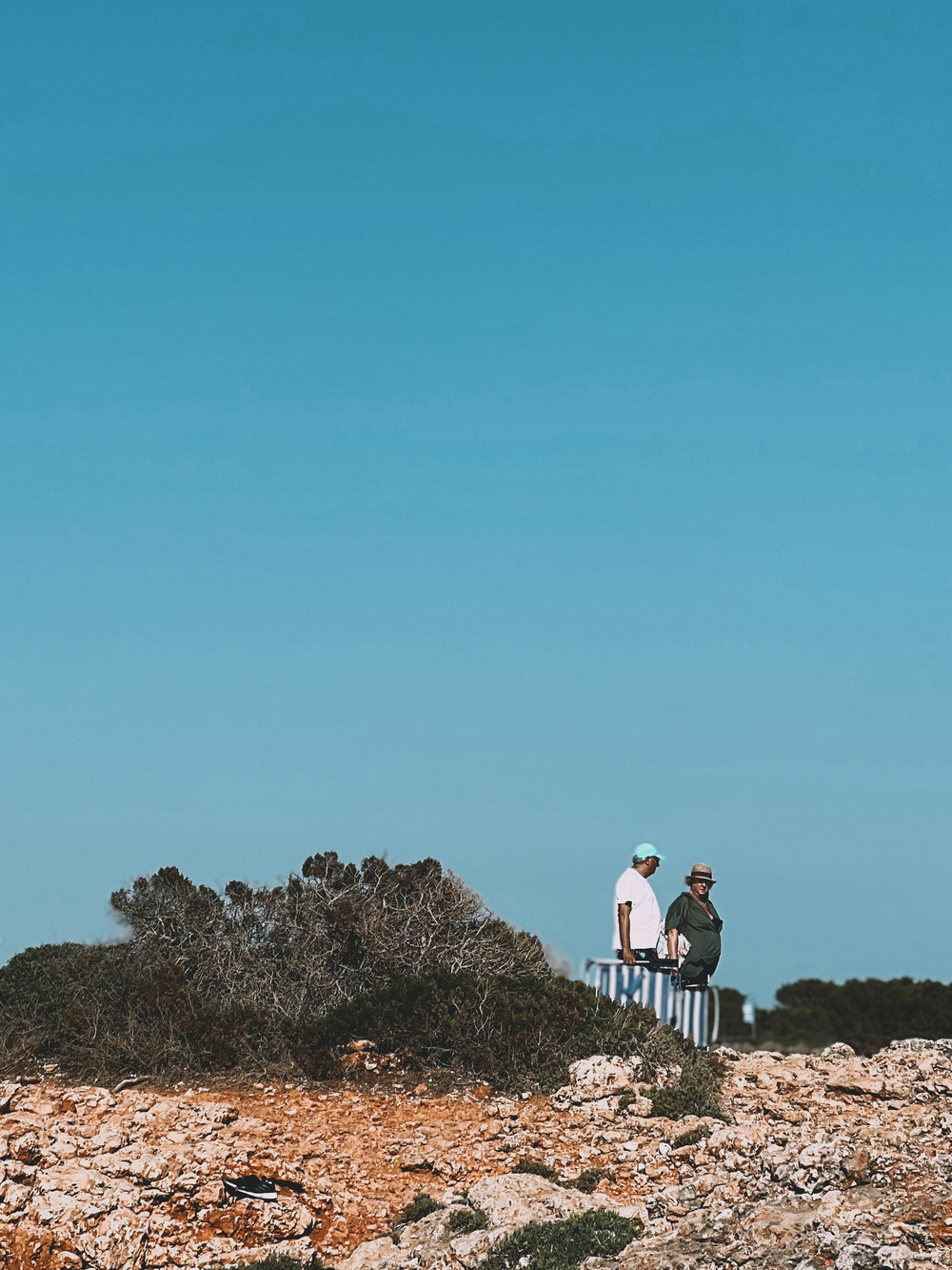 A couple stands on a rocky and arid landscape with some shrubs, under a clear blue sky. They are wearing casual clothing and hats and are looking towards the horizon.