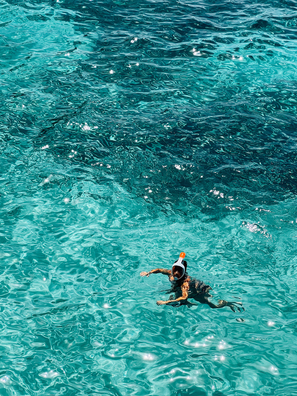 A person snorkeling in clear, turquoise water with ripples on the surface. The snorkeler is wearing a full-face snorkel mask with an orange top.