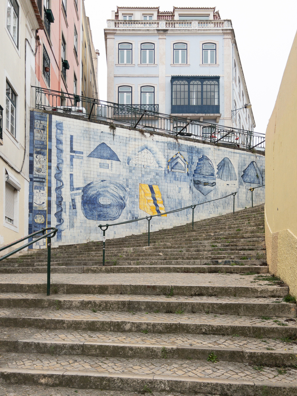 The image shows a staircase in an urban area with a hand-painted mural on the adjoining wall. The mural features abstract blue and yellow designs. Tall buildings with multiple windows and pastel-colored facades surround the staircase.