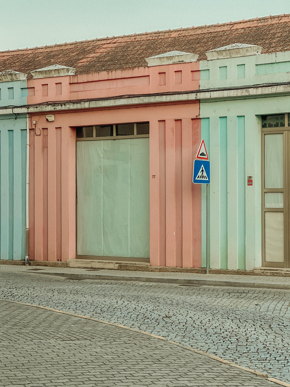 A pastel-colored building with sections painted in light pink, light blue, and light green. It features vertically grooved columns and a red-tiled roof. In front of the building, there is a pedestrian crossing sign and a cobblestone street.