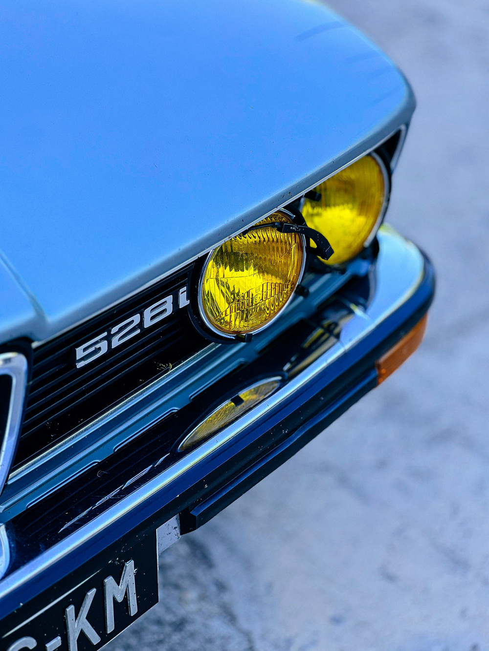 Close-up of the front of a vintage blue BMW 528i, featuring distinctive yellow headlights and part of a black license plate with silver characters.