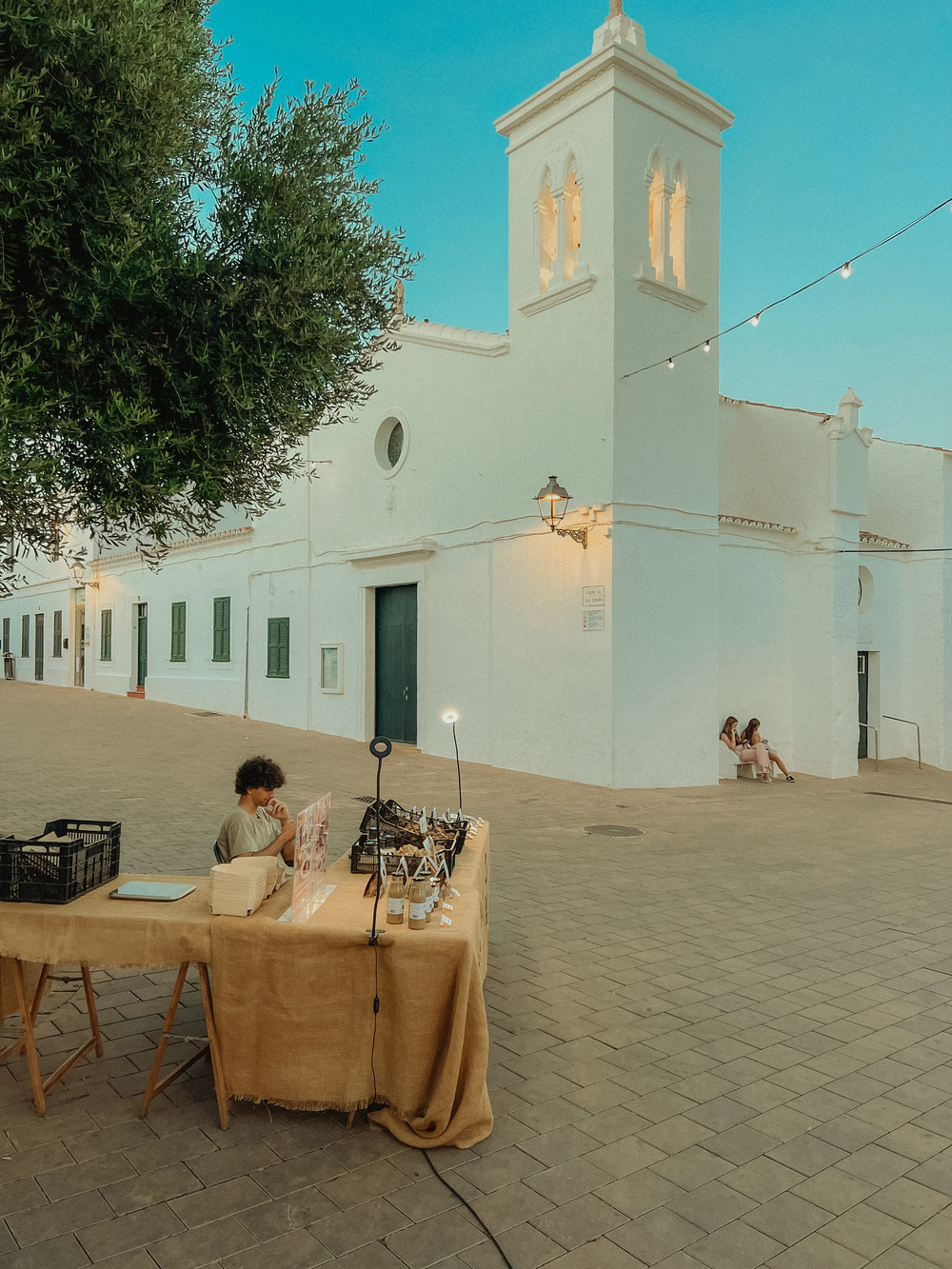 A street vendor table is set up with various items for sale, including bottles and display stands, with a person sitting behind it. The table is covered with a beige cloth. In the background, there is a white church with green doors and windows.