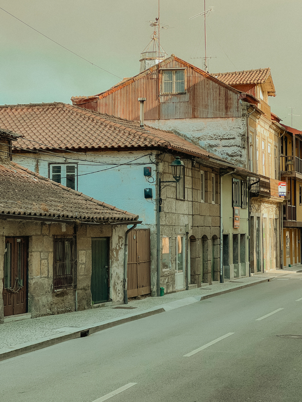 A narrow street lined with old houses, featuring stone and plaster facades. The buildings have tiled roofs with visible wear and several television antennas on top. The street is empty, with a cobbled sidewalk and a lantern attached to one of the buildings.