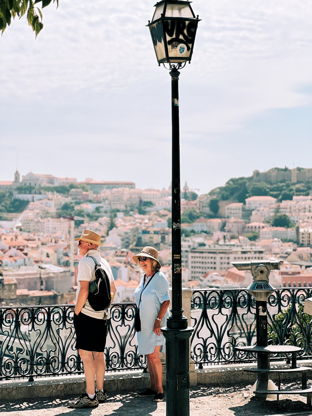 A man and a woman stand at a scenic viewpoint overlooking a city, separated by a vintage-style lamppost with decals. The man wears a hat, sunglasses, a white shirt, black shorts, and a backpack, while the woman wears a hat, and a dress.