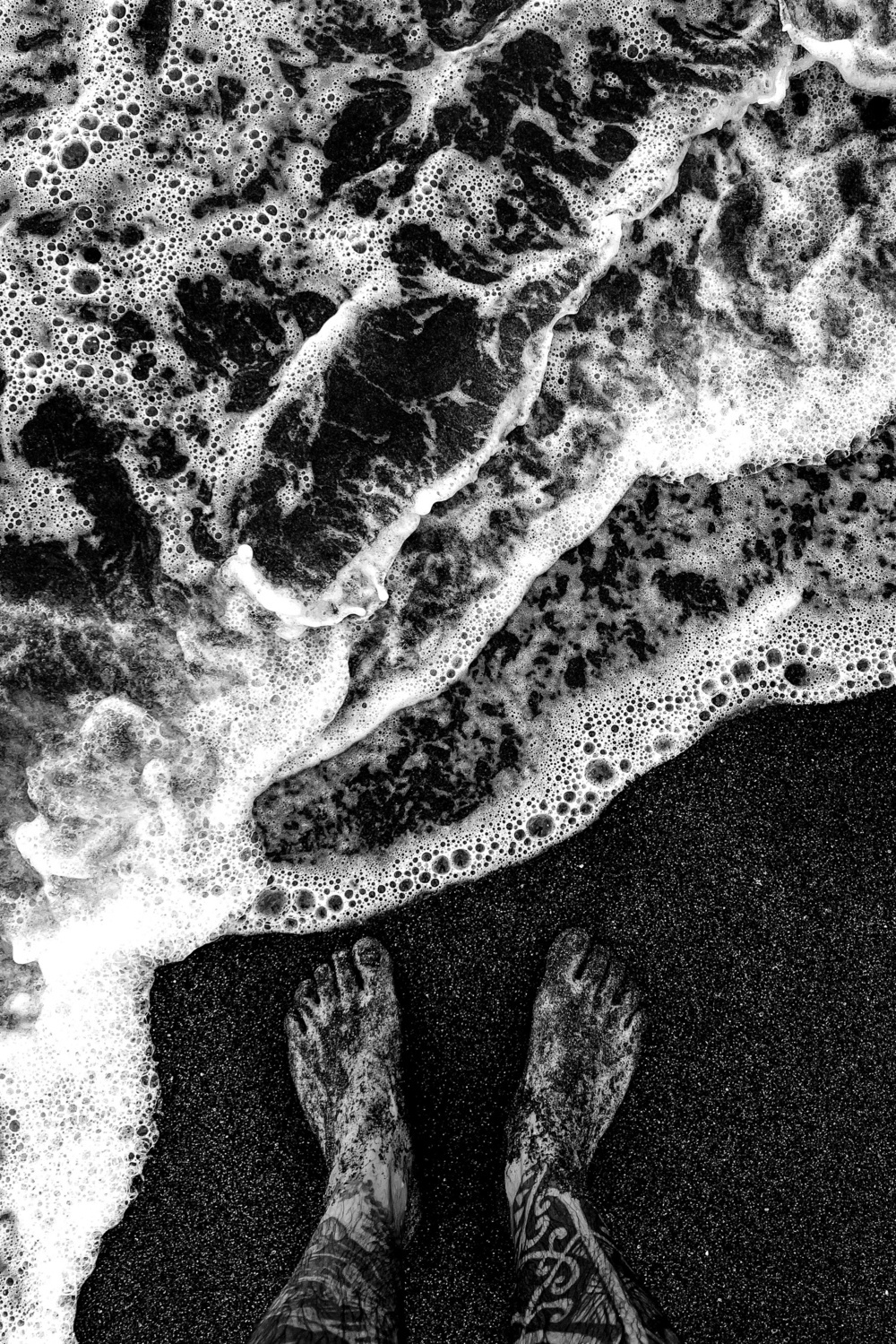 A black-and-white photo of sandy feet with tattoos standing on a shoreline as waves from the sea foam around them.