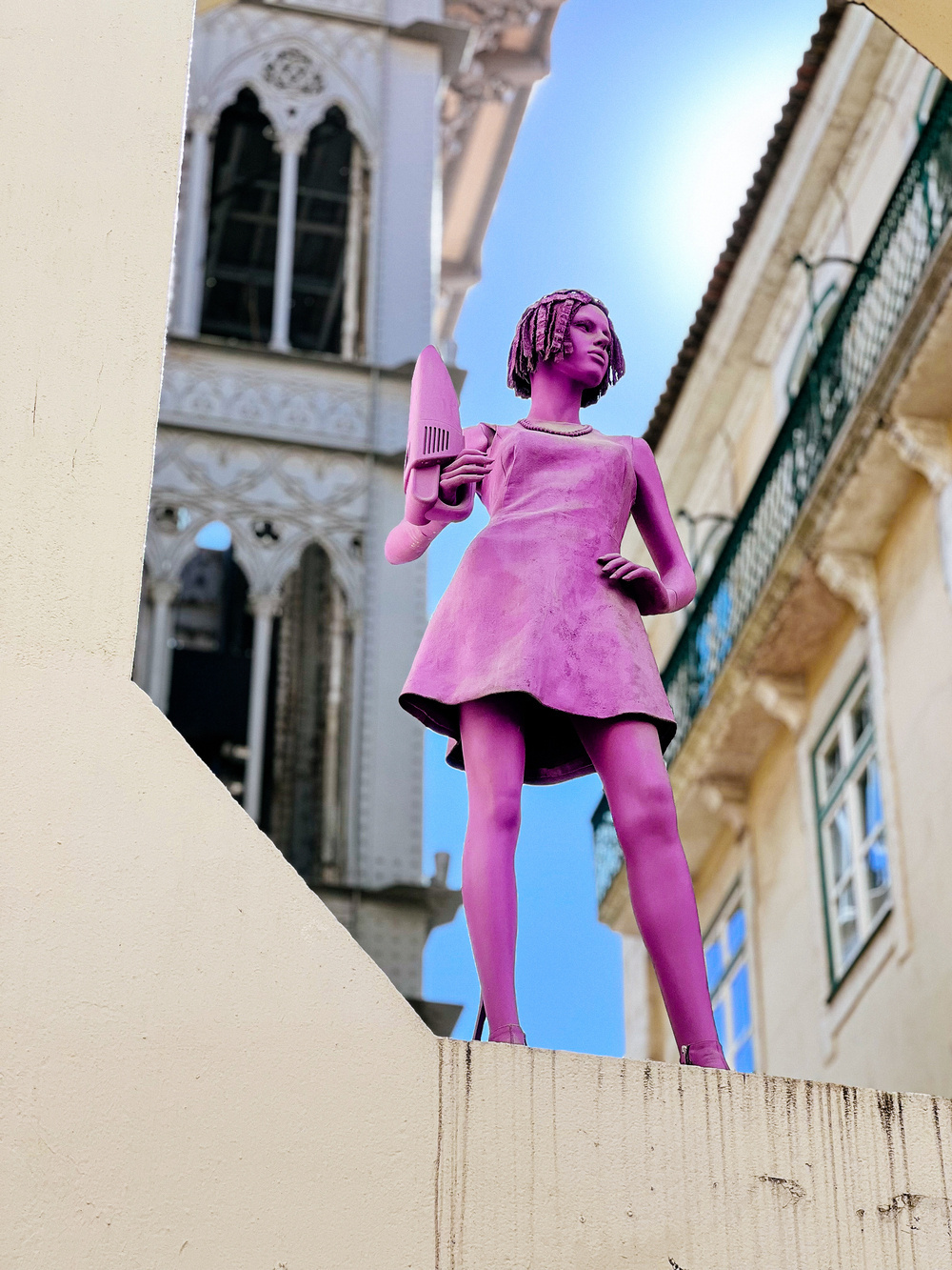 A pink statue of a woman holding a hairdryer stands on a ledge against the backdrop of a historic building with ornate windows and a clear blue sky.