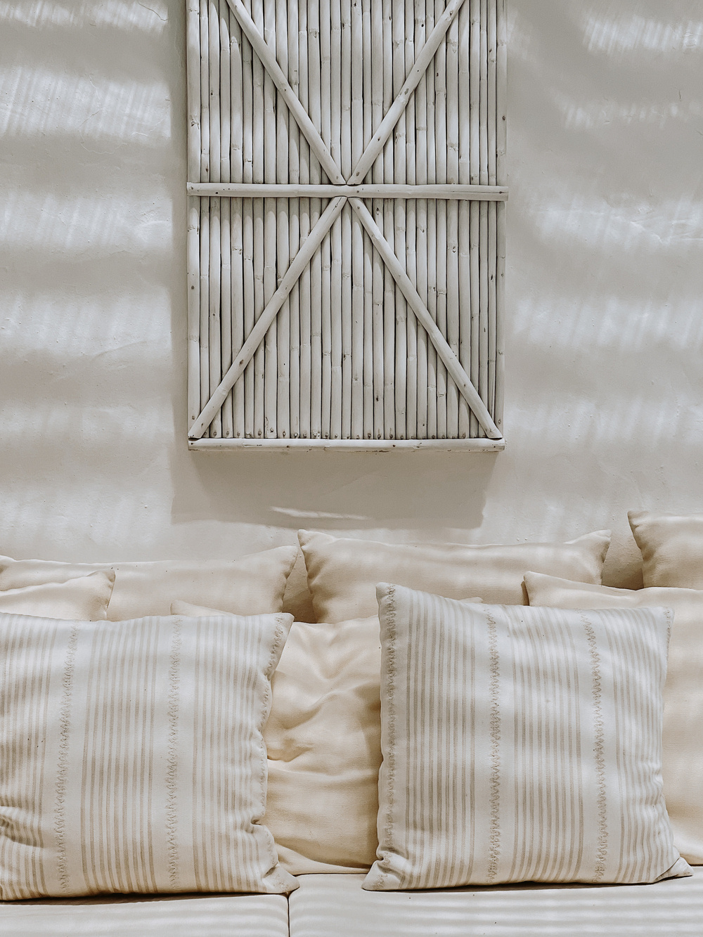 Decorative wicker wall art with an &ldquo;X&rdquo; pattern hangs above a cream-colored sofa adorned with several beige and white striped throw pillows. The room has a serene and minimalist aesthetic.