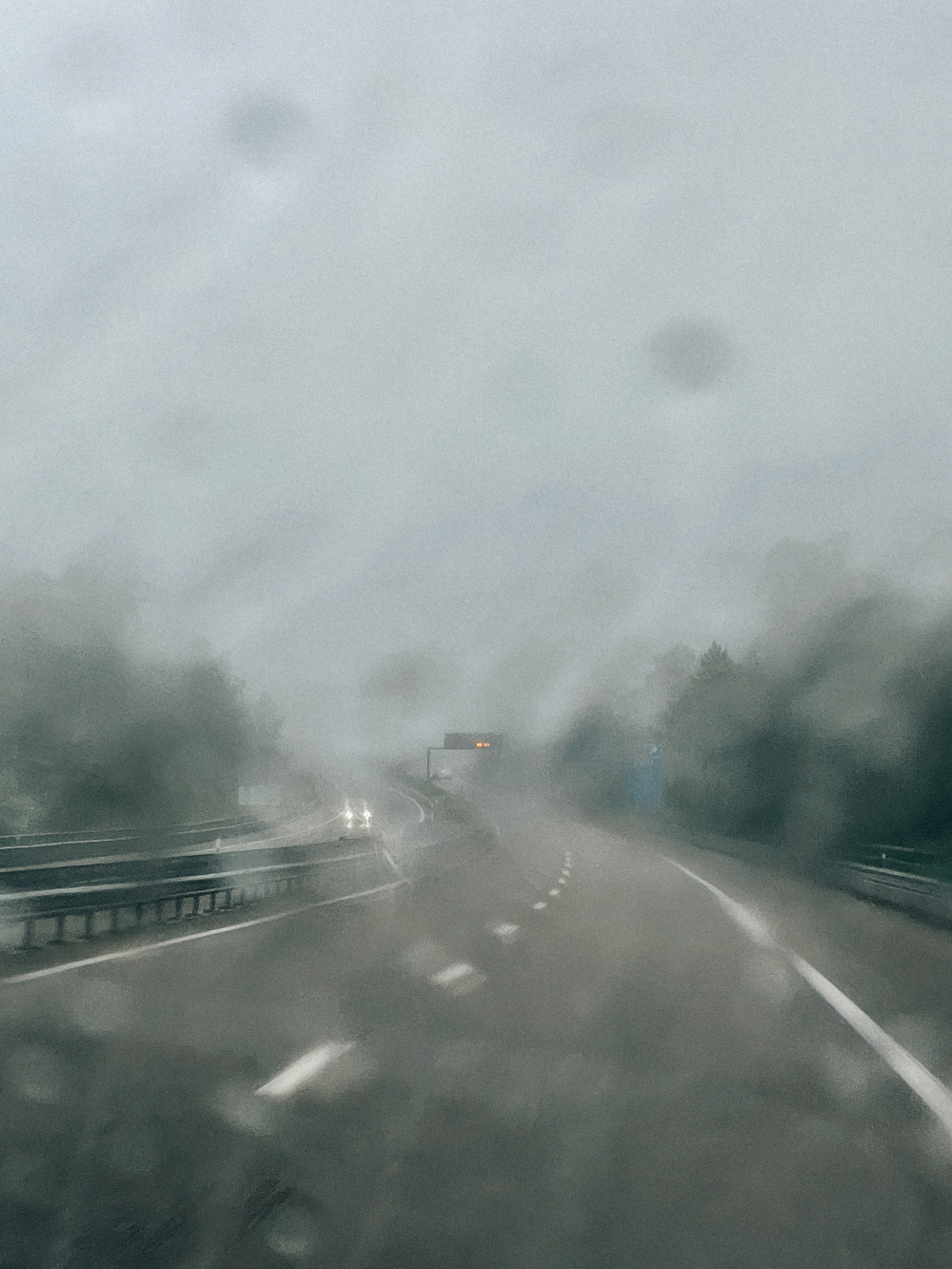 A blurred view of a highway on a rainy, foggy day, with water droplets on the camera lens. The road is mostly empty, with only a few cars visible, some with headlights on. The surroundings are obscured due to the weather conditions.