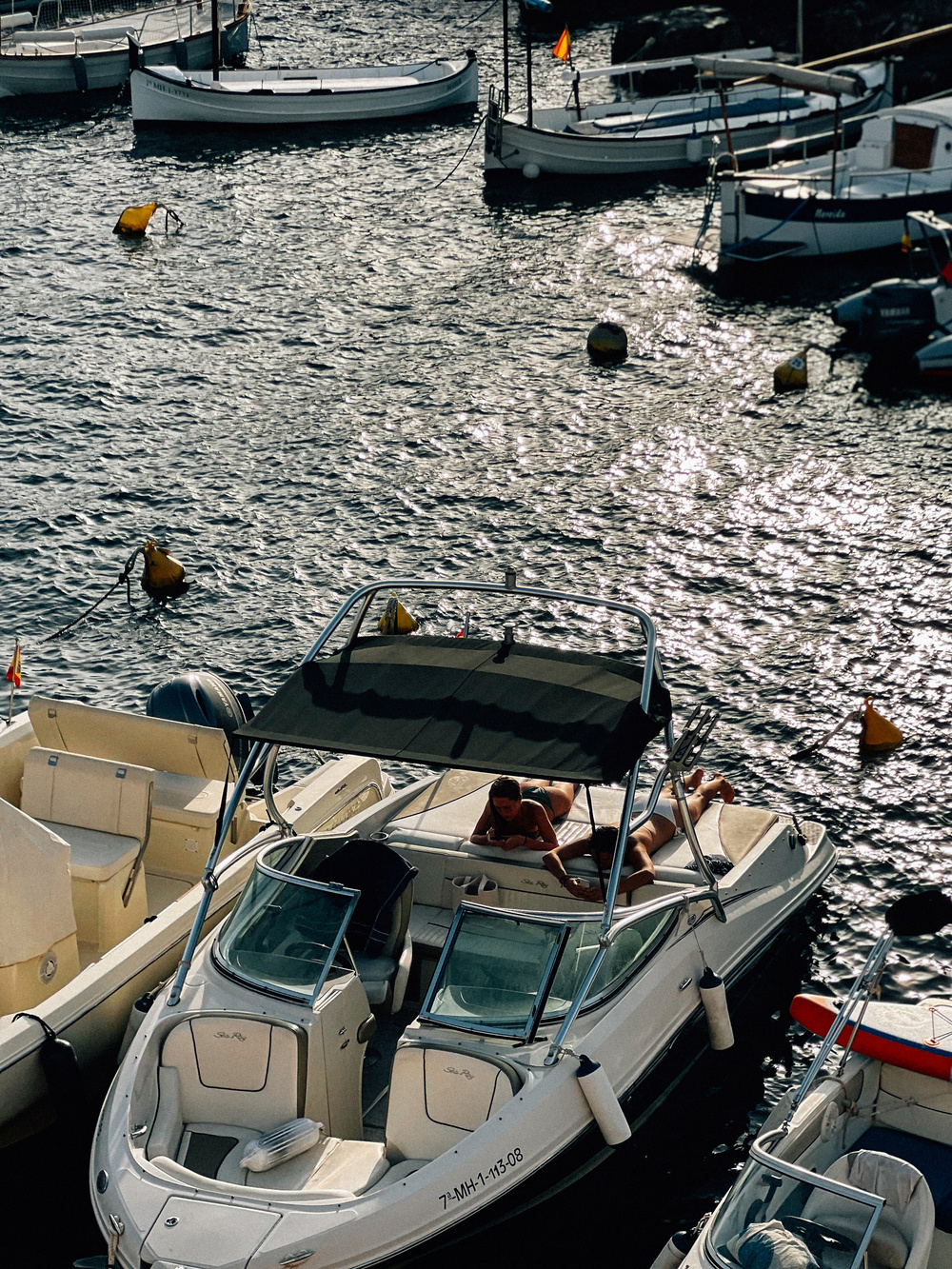 A small marina with several docked boats, including a white motorboat in the foreground. Women are sunbathing on the deck of the motorboat. The water is calm, and sunlight reflects off its surface.