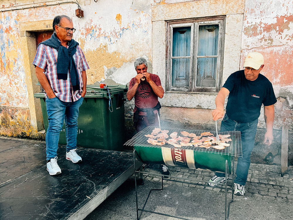 Three men outdoors near a building with peeling paint; one grilling meat, another watching, and the third drinking from a small cup.