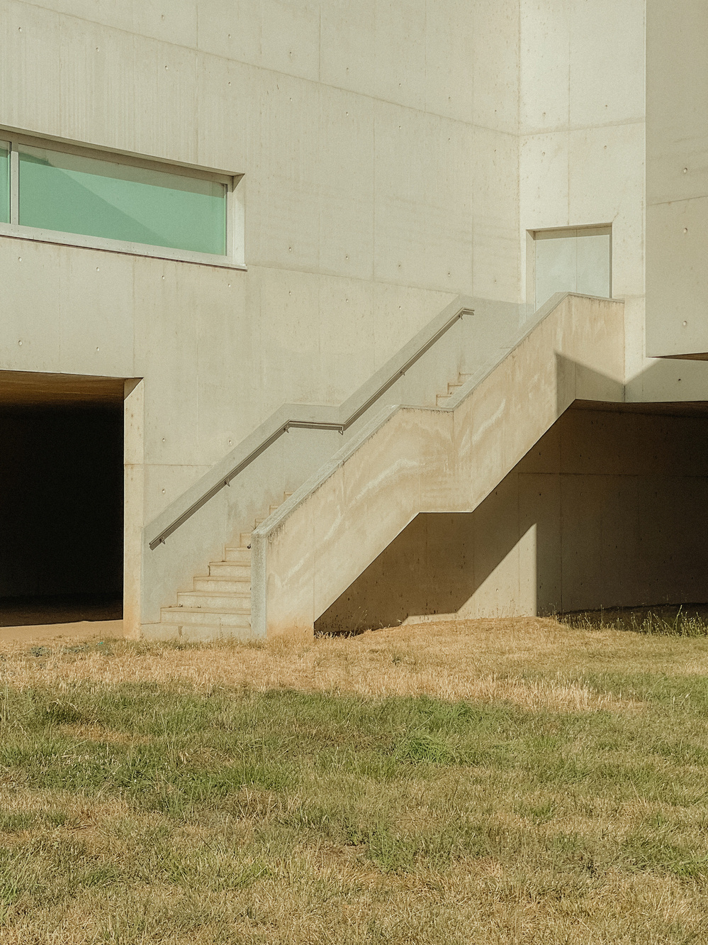 Outdoor concrete staircase attached to a building, with minimalistic architectural design. The structure includes rectangular windows and a grassy area in the foreground.