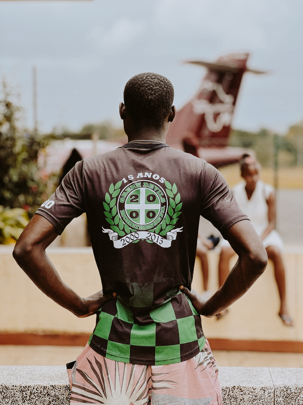 A person wearing a black and green shirt with the text “15 ANOS,” “2000-2015,” and a crest logo standing with hands on hips. A blurred background shows part of an aircraft and another person sitting.