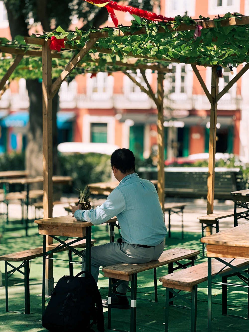 A man in a light blue shirt sits alone at a wooden picnic table under a pergola adorned with green vines and red ribbons. His black backpack is on the ground next to him, and he appears to be holding a pineapple.