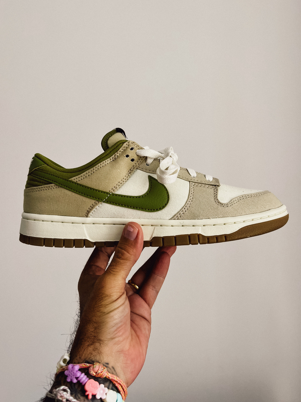 A hand is holding a beige and green Nike sneaker with white laces against a plain background. The hand is adorned with colorful bracelets and a ring.