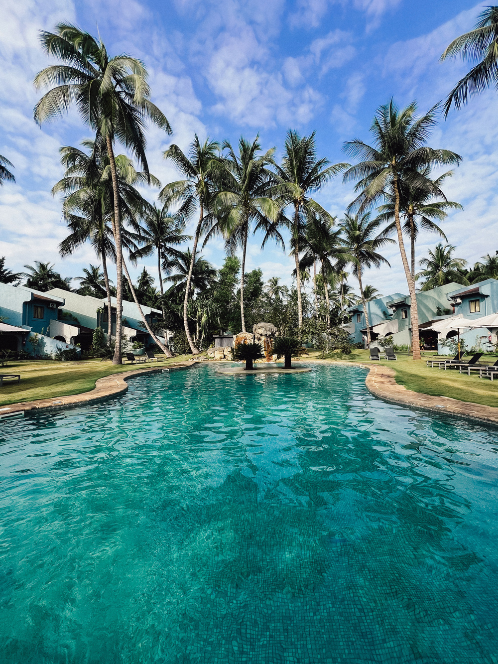 A serene outdoor swimming pool surrounded by lush palm trees and neat green lawns. In the background, there are bungalow-style buildings under a partly cloudy blue sky. The area exudes a tropical, vacation-like atmosphere.