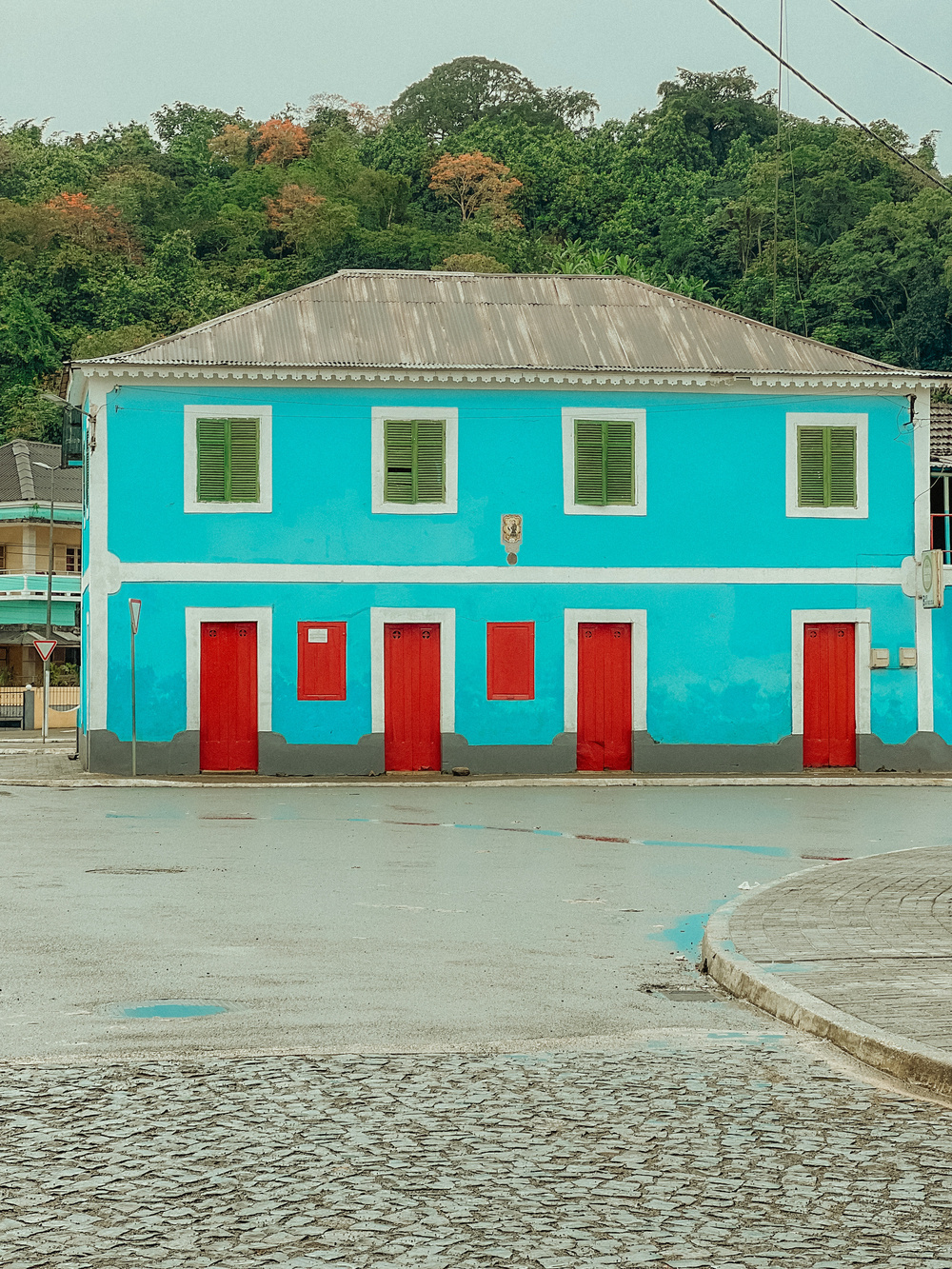 A vibrantly painted building with turquoise walls, green shutters, and red doors stands on a slightly wet cobblestone street. The structure sits against a lush, green, forested background.