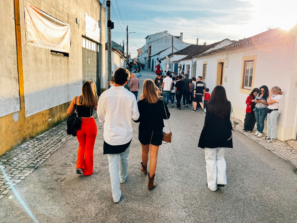 A group of people walking down a street in a town, potentially heading towards an event with other attendees visible ahead. The sun is low in the sky, creating long shadows, and the architecture suggests a European setting.