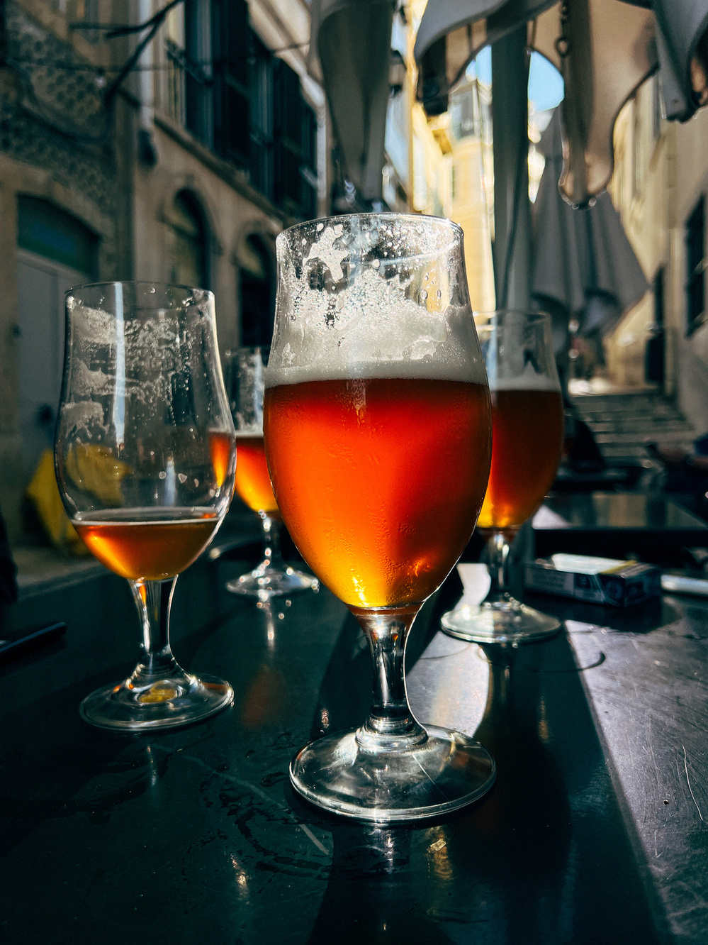 Four glasses of amber-colored beer on a dark table outdoors, in a narrow, sunlit alley, with buildings in the background.