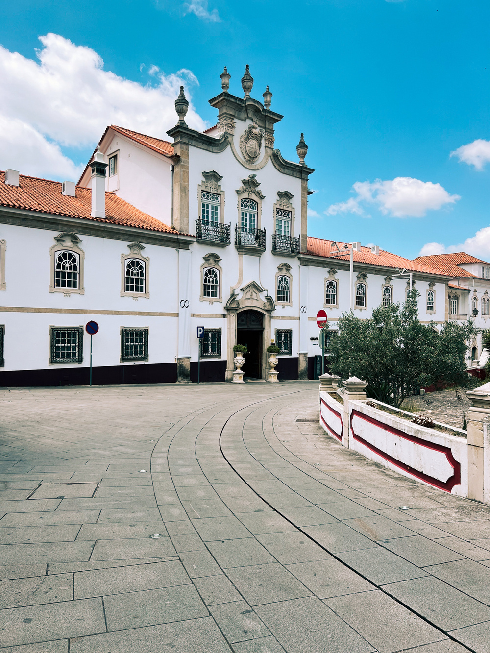 A historic building with white-washed walls, ornate architectural details, and a red-tiled roof under a blue sky with scattered clouds. The building features arched windows, a central entrance with decorative elements, and a balcony.