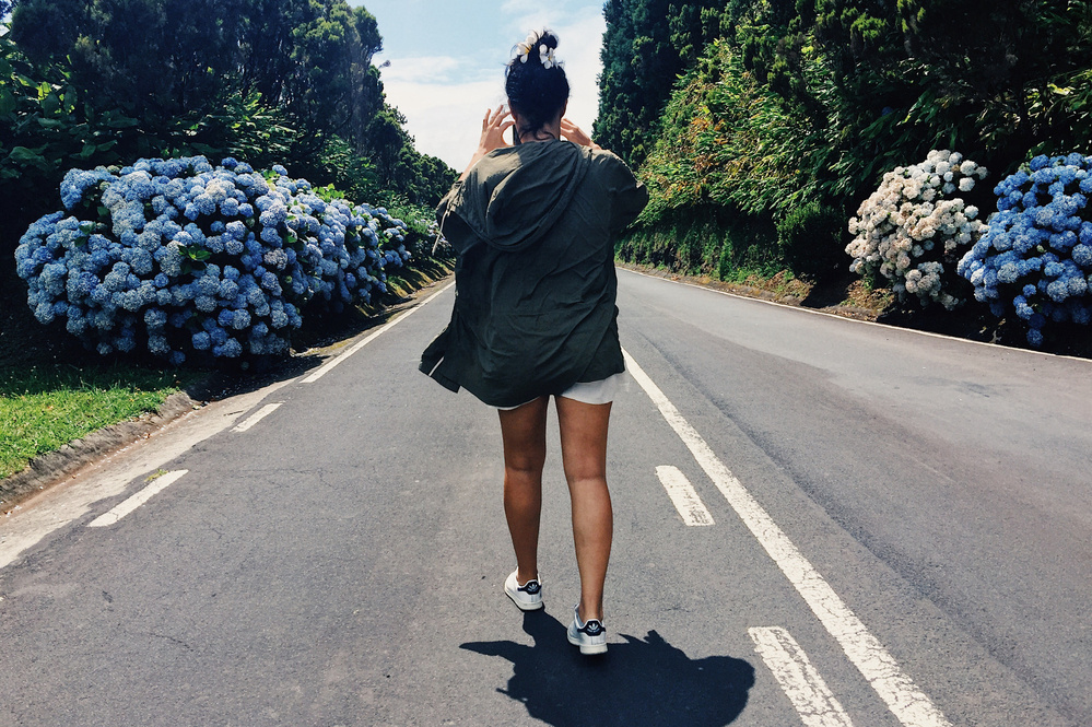 A person is walking down the middle of an empty road surrounded by lush greenery and large, vibrant hydrangea bushes on both sides. They are wearing a green jacket, white shorts, and white sneakers, with their back to the camera. 