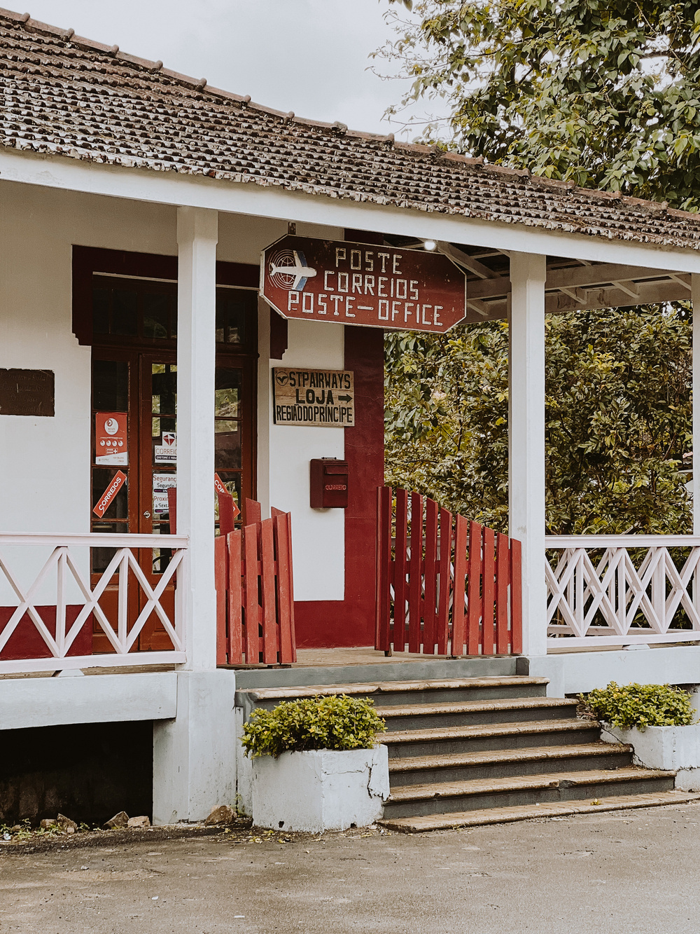 A small, rustic post office with a tiled roof and red and white painted exterior. The building has steps leading up to the entrance and a wooden sign displaying “Posto Correios Poste Office.” There are plants next to the steps and greenery surrounding the building. 