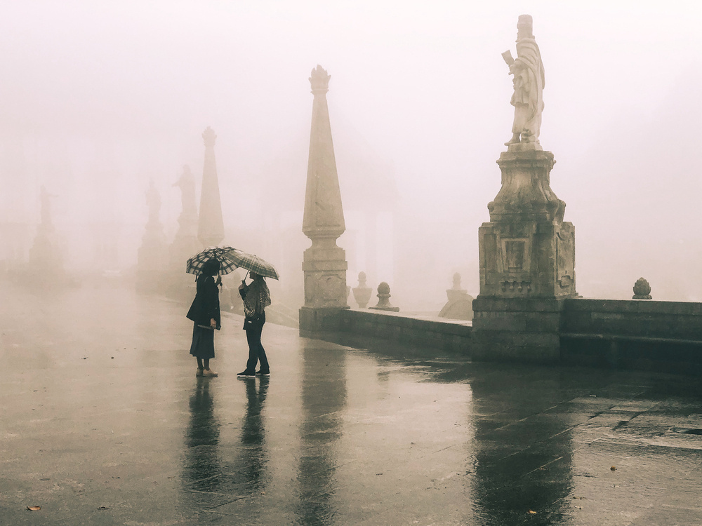 Two people standing under an umbrella on a foggy day with historic statues and wet ground visible in the background.