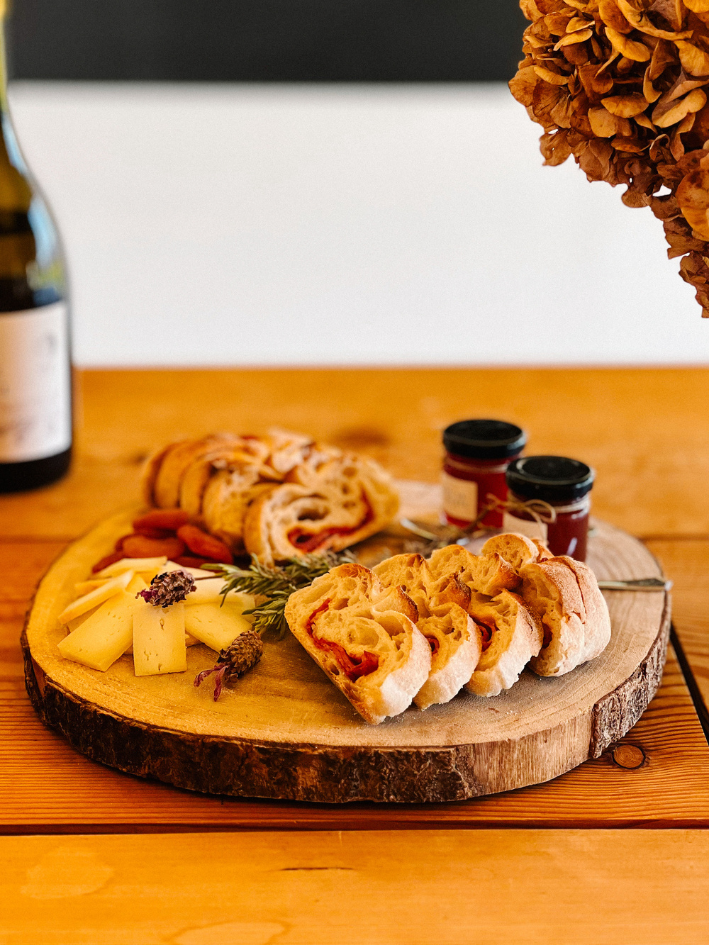 A cheese platter with sliced baguette, cheese, and condiments on a wooden board, with a bottle of wine and flowers in the background.