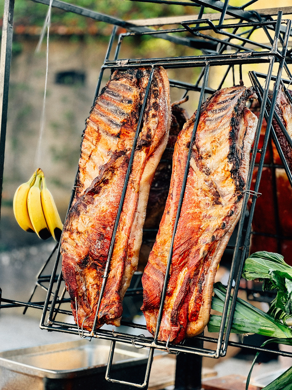 Grilled pork ribs hanging on a large outdoor rack with a bunch of bananas and green leaves in the background.