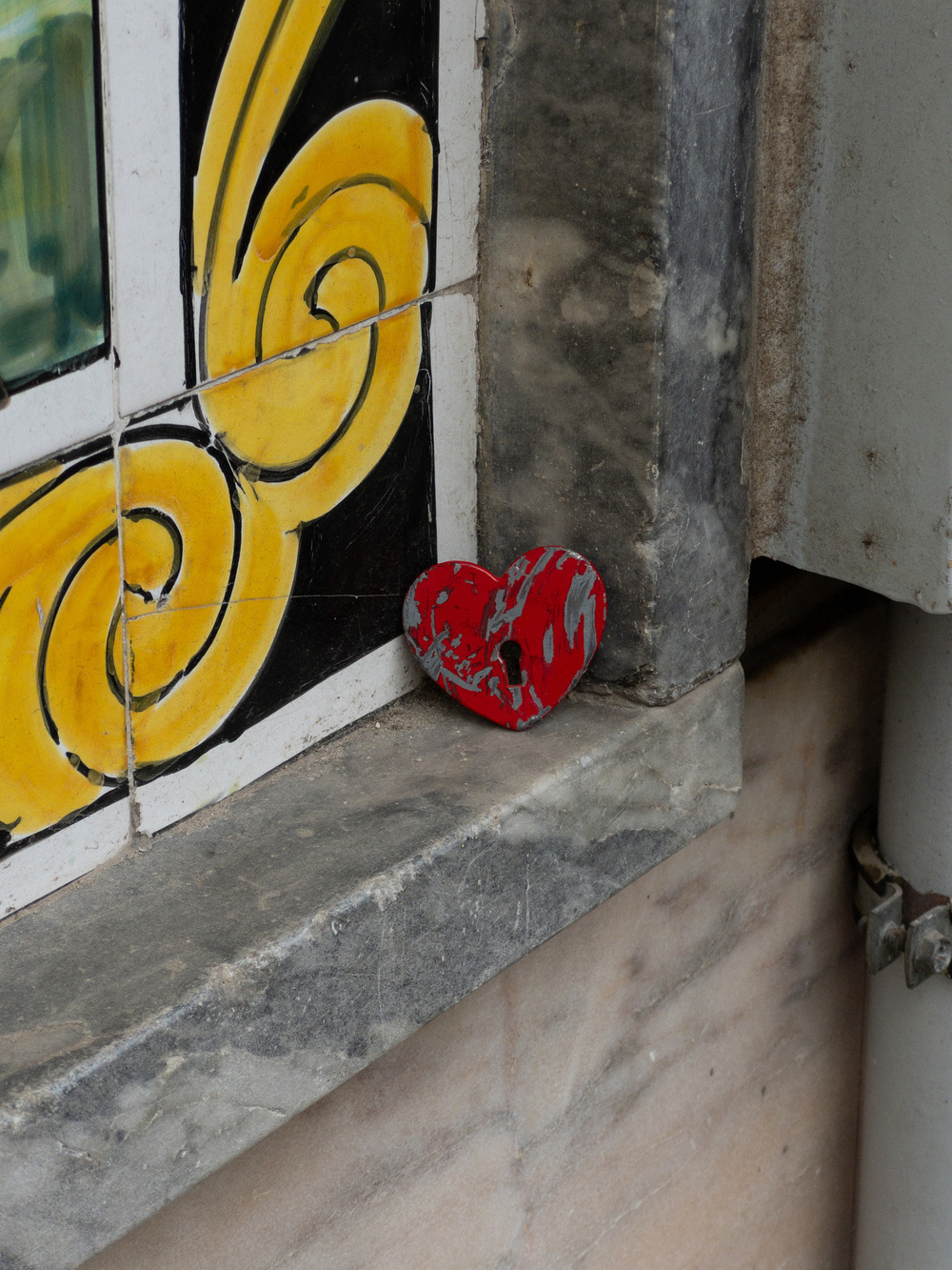 A small, painted red heart shape is placed on a ledge next to a decorative tile with yellow and black scrollwork. The ledge is made of stone or marble, showing some wear and tear.