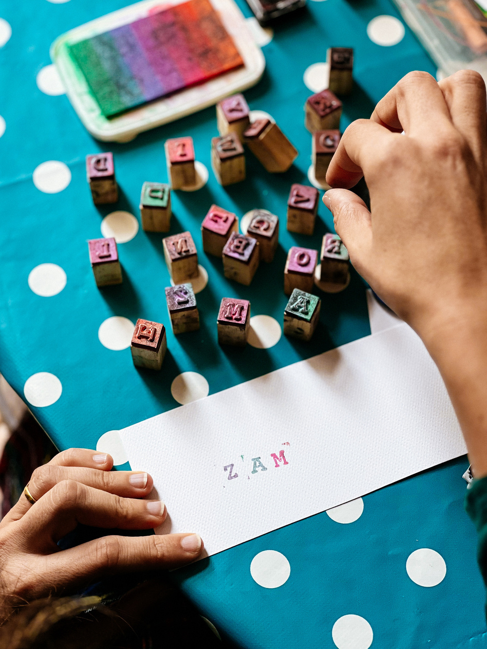 A person&rsquo;s hands stamping a card with colored alphabet stamps and an ink pad on a polka-dot tablecloth.