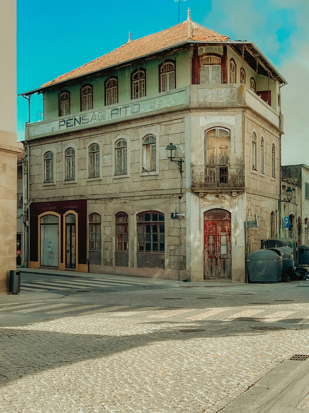 A vintage, weathered three-story building with faded signage &ldquo;Pensão Rito&rdquo; is seen on a cobblestone street corner. The ground floor has boarded-up windows and doors, while the upper floors feature multiple arched windows.