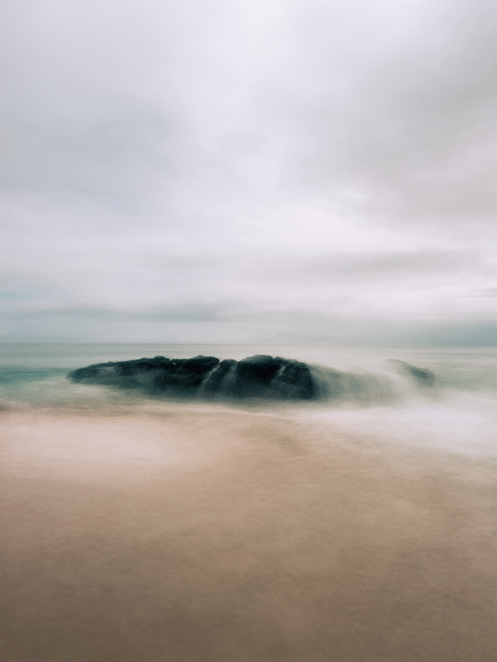 A serene coastal scene featuring a rocky formation partially submerged in the ocean, with misty waves gently crashing against it. The sky is overcast, creating a soft, muted atmosphere.