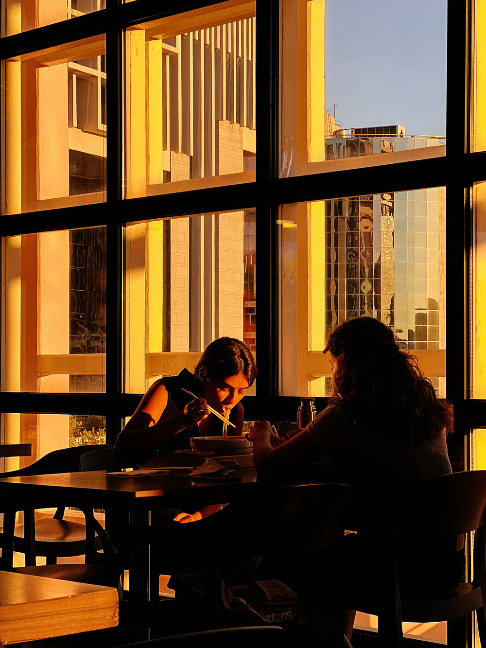 Two individuals are dining at a table inside a restaurant with large windows. One of them is using chopsticks to eat. The scene is bathed in the warm light of the golden hour, with a cityscape visible outside the windows.