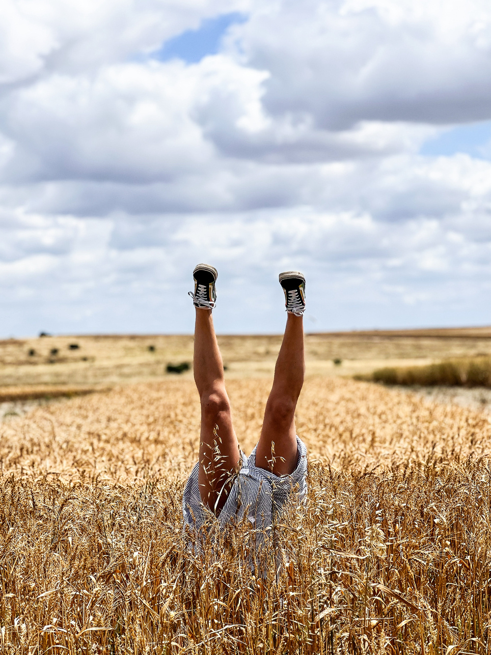 The image shows a person upside down in a wheat field, with only their legs and feet visible. They are wearing black sneakers and shorts. The sky is cloudy with a blue patch visible.