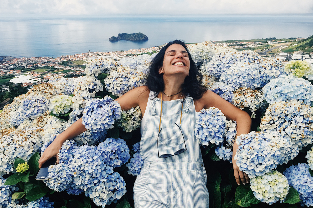 A woman stands joyfully amidst a large cluster of blue and white hydrangeas, with a view of the coastline and ocean in the background. She is wearing a striped overall and has a big smile on her face. The scene is bright and picturesque.