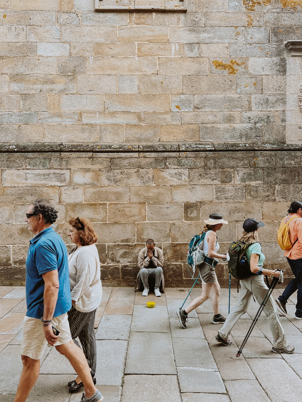 A person sits against a brick wall with a yellow bowl on the ground in front of them, appearing to ask for help or donations. Several other individuals, some with walking sticks and backpacks, pass by without engaging.